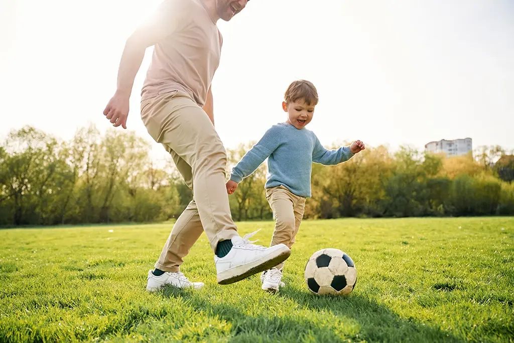 A father and son are running and playing soccer together in a field outside.