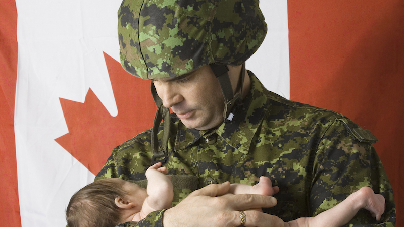A soldier holds his tiny baby, as though leaving for combat duty, with the Canadian flag as a backdrop.