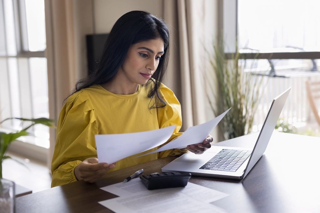 Woman wearing yellow blouse sits at table with laptop, reviewing insurance documents.