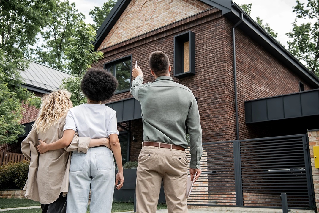 A couple with arms around stand next to a man who is pointing to a brick house.