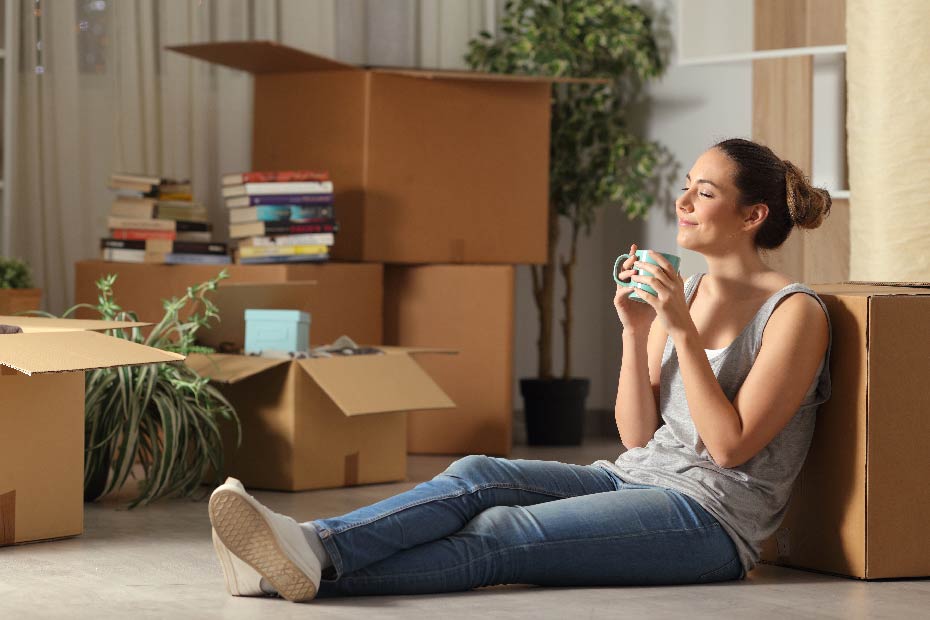 Woman sitting down and drinking tea beside open cardboard boxes.